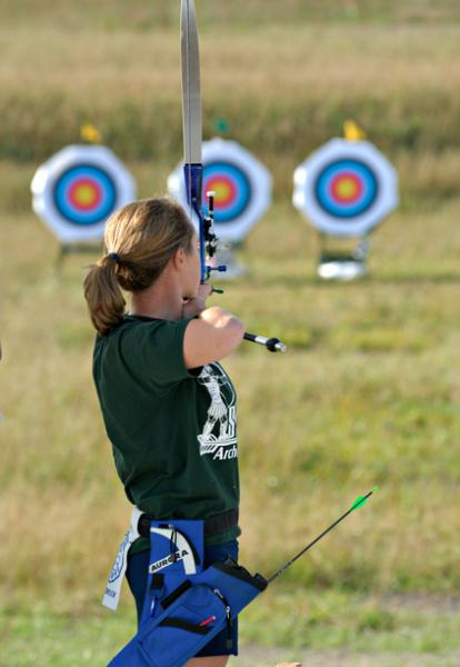 A young girl practicing archery outdoors with three target boards in the background.