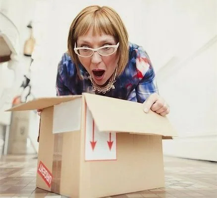A woman with glasses and short hair looks surprised as she peers into a cardboard box on the floor.