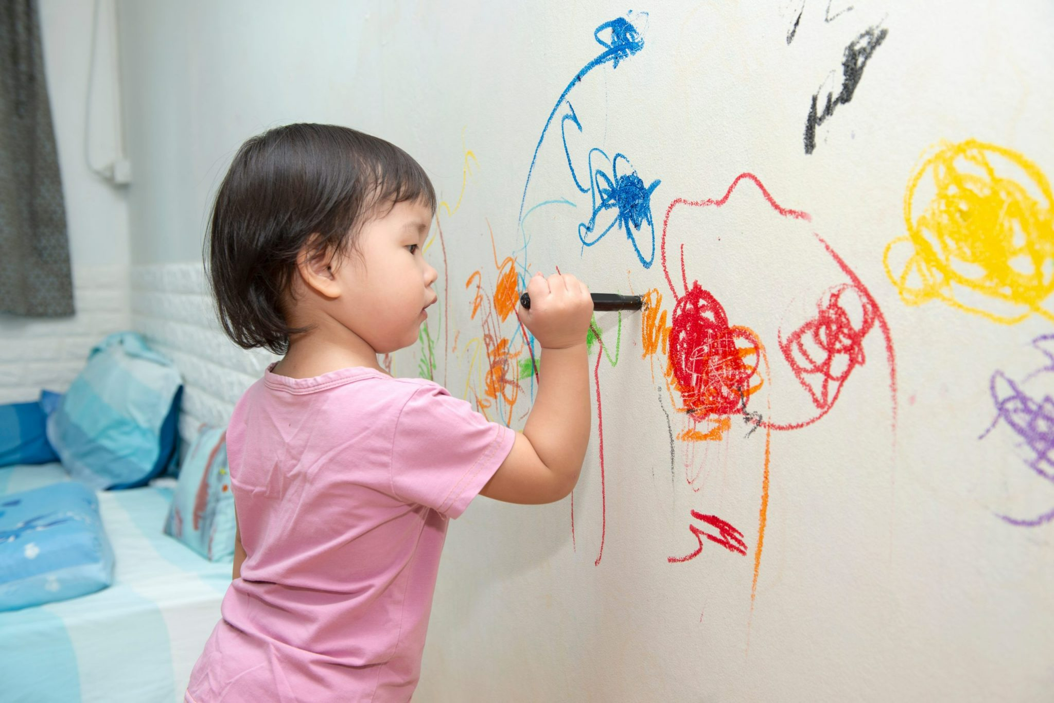 A young girl in a pink shirt is drawing with a black marker on a white wall, creating colorful scribbles and doodles.